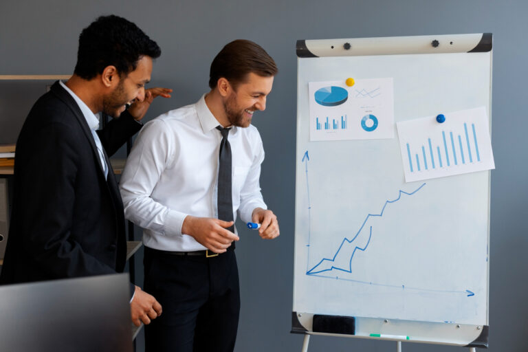 Two business men strategizing together next to a whiteboard that features graphs relevant to their business discussions.